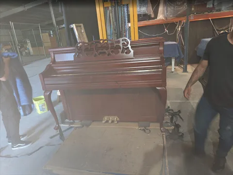 Upright piano on wheeled support beside shelving and wrapped instruments